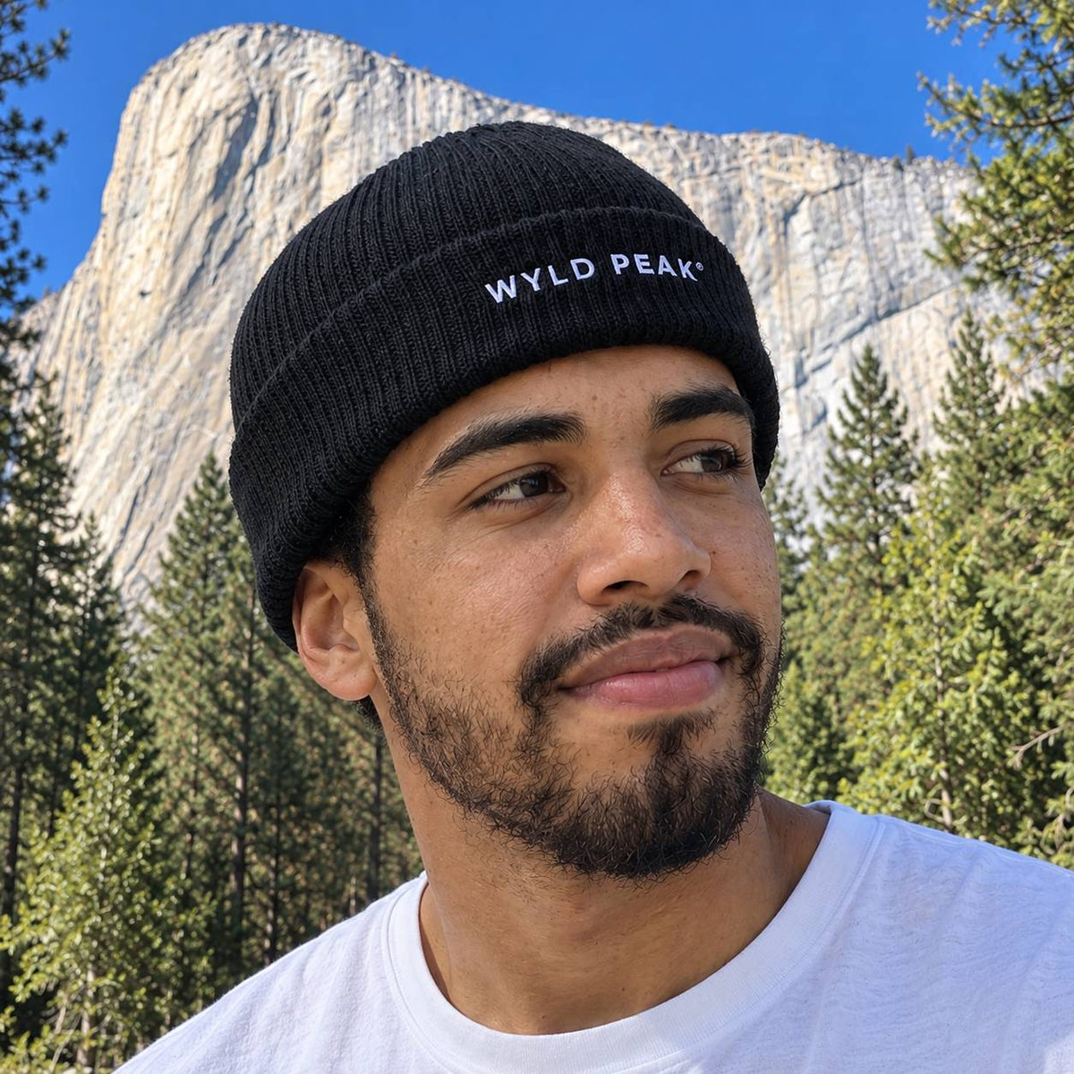 A man wearing a black knit beanie with 'WYLD PEAK' embroidered in white on the front, photographed outdoors in front of a large rock formation and trees under a blue sky.