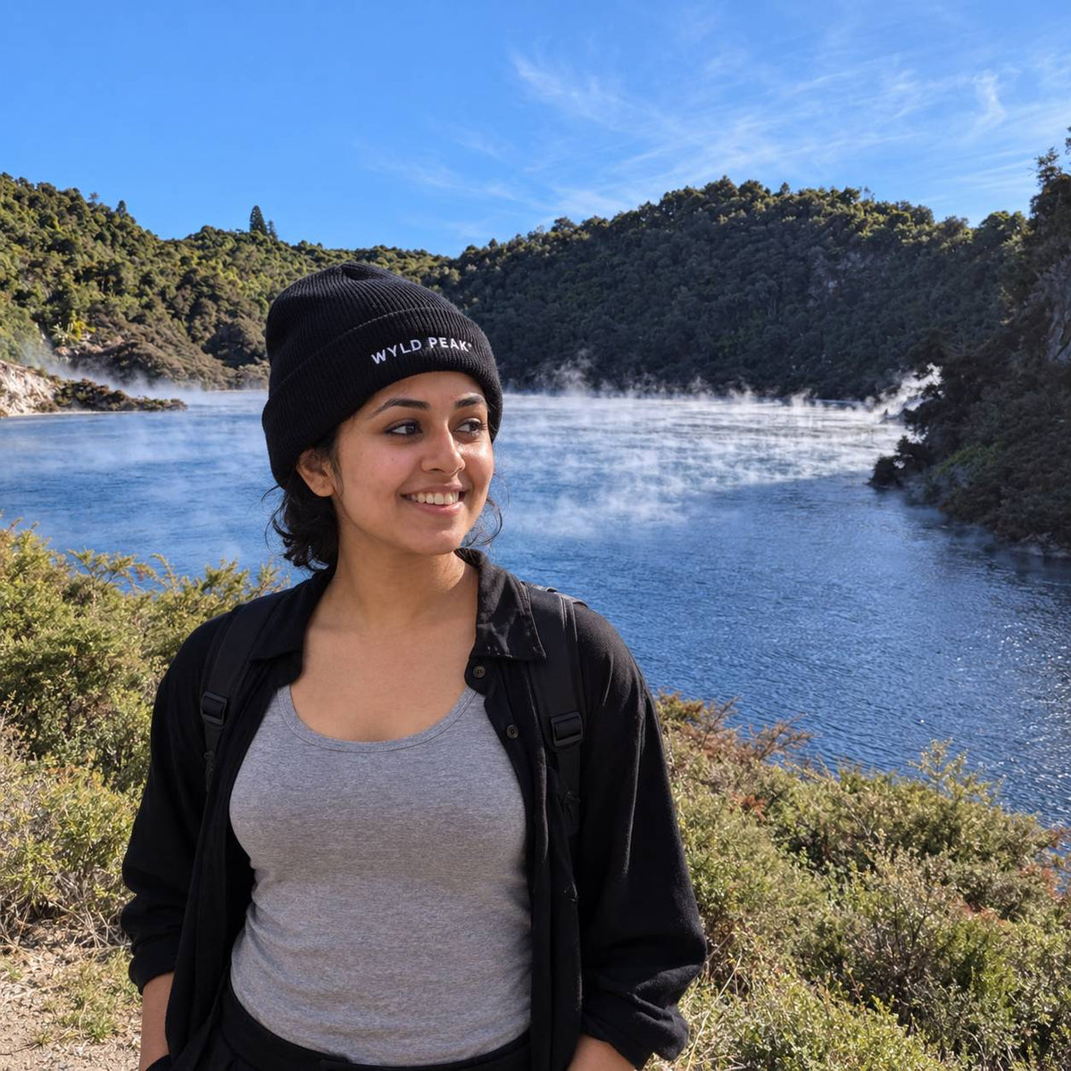 A woman wearing a black beanie with 'WYLD PEAK' embroidered on the front, a black jacket, and a gray tank top stands outdoors in front of a scenic lake and forested hills under a blue sky.