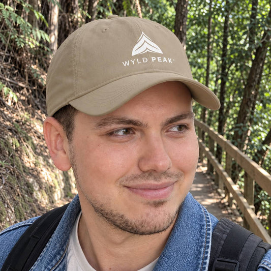 A man wearing a tan baseball cap with a white 'WYLD PEAK' logo, photographed outdoors on a wooded path with a wooden railing.