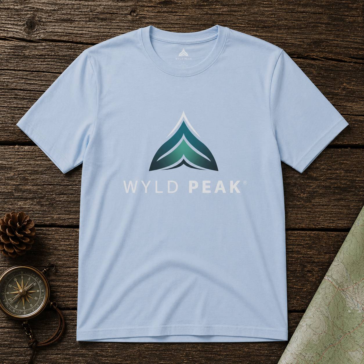 Light blue t-shirt with 'WYLD PEAK' logo on a wooden surface with nature-themed items.