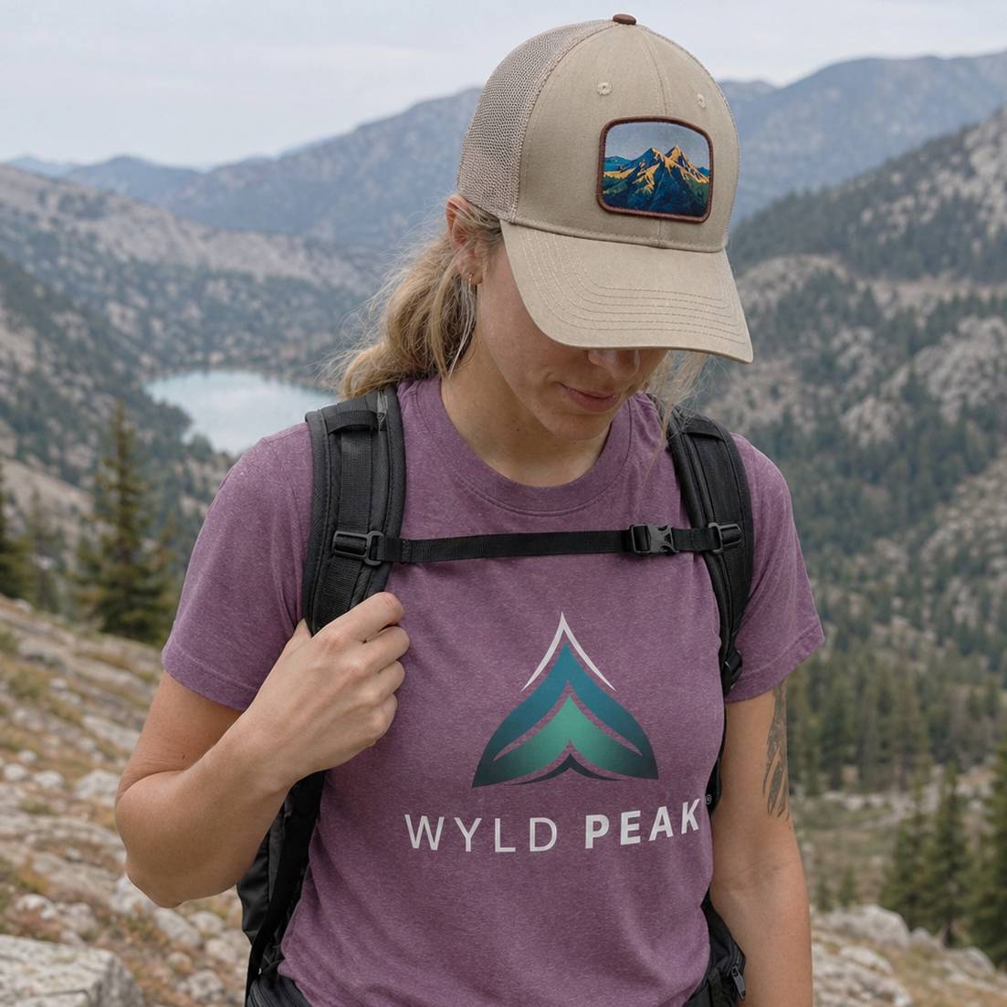 Person wearing a purple 'Wyld Peak' t-shirt and beige cap with mountain design, hiking in a scenic mountainous area.