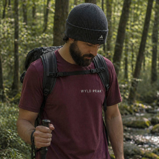 Man hiking in a forest wearing a maroon 'Wyld Peak' t-shirt and black Adidas beanie.