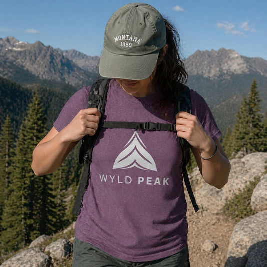 Person wearing a purple 'Wyld Peak' t-shirt and green cap with mountains in the background