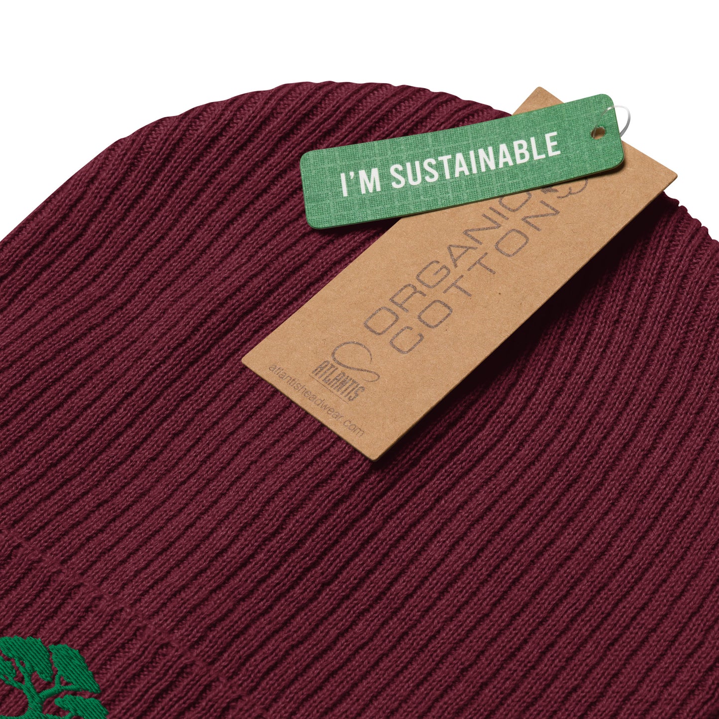 A close-up of a ribbed knit beanie in deep burgundy, with a green 'I'M SUSTAINABLE' tag and a brown paper cotton tag attached. The background is solid white.