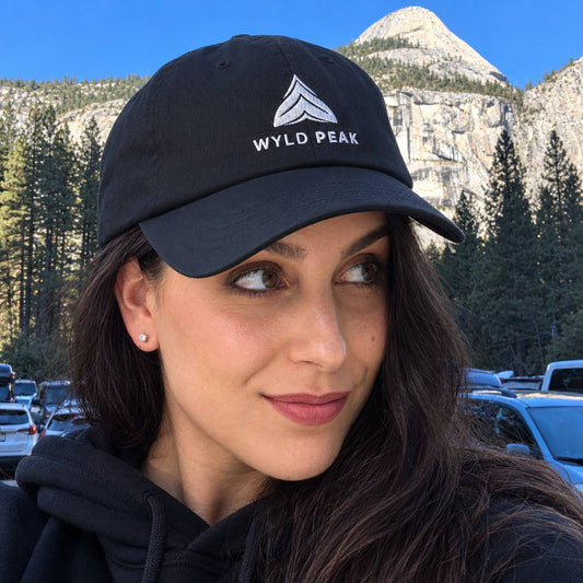 A woman wearing a black baseball cap with a white geometric logo and the text 'WYLD PEAK' in front of a mountain and forest background.