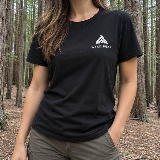 A woman wearing a black short-sleeve t-shirt with a white Wyld Peak logo on the left chest, standing in a forest.