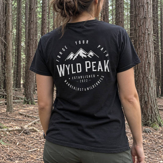A woman viewed from behind wearing a black t-shirt with a white graphic print that reads 'WYLD PEAK' and 'FORGE YOUR PATH' in a forest setting.