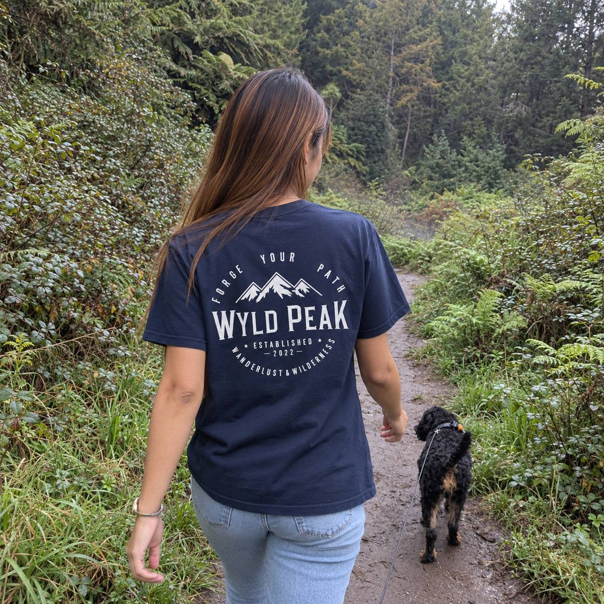A woman wearing a navy blue t-shirt with a white 'WYLD PEAK' mountain graphic on the back, walking on a forest path with her dog.