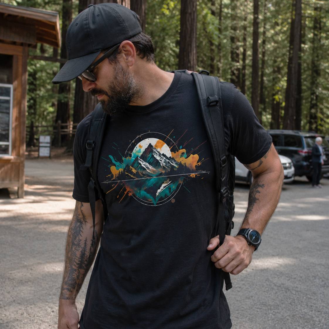 Man wearing a black t-shirt with a mountain graphic and a backpack, standing in a forested area.