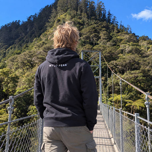 A person viewed from behind wearing a black hoodie with 'WYLD PEAK' printed in white on the back, standing on a suspension bridge overlooking a lush green forested mountain under a blue sky.