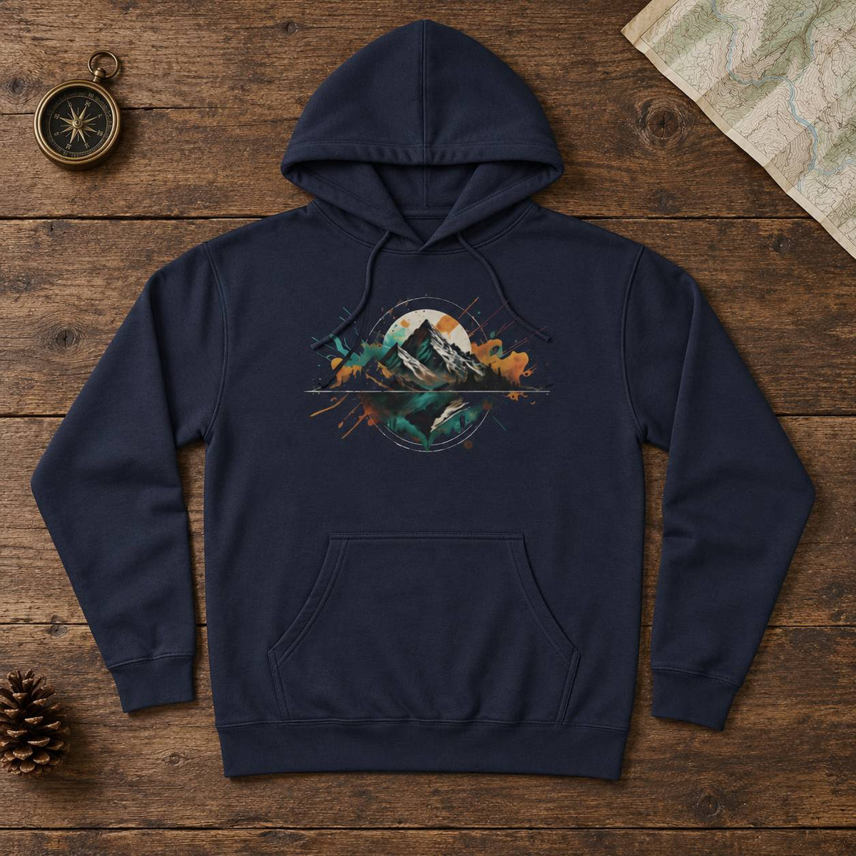 A navy blue pullover hoodie with a colorful mountain and lake graphic on the chest, laid flat on a rustic wooden surface with a compass, map, and pinecone nearby.