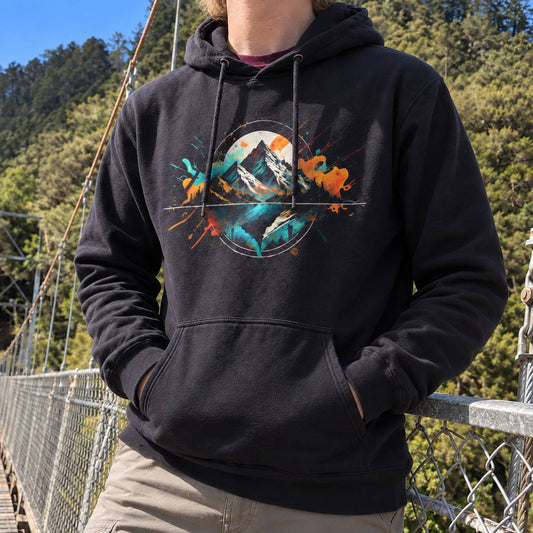 A person wearing a black pullover hoodie with a colorful mountain graphic on the chest, standing on a suspension bridge with trees in the background.