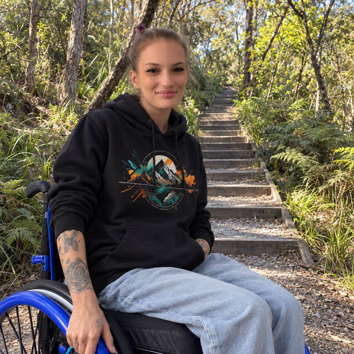 A woman with blonde hair tied back sits in a blue wheelchair on a forest hiking trail, wearing a black hoodie with a colorful mountain and bird graphic on the chest, paired with light-wash jeans.