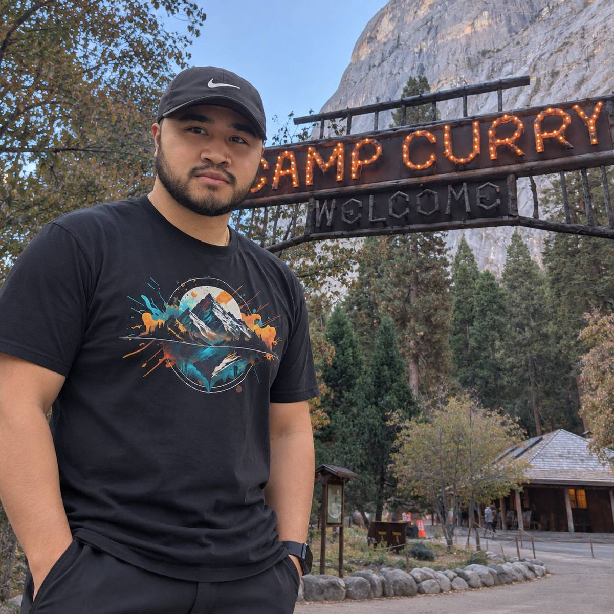 A man wearing a black Nike cap and a black graphic t-shirt stands in front of a 'Camp Curry Welcome' sign and a large rock cliff, likely in Yosemite National Park.