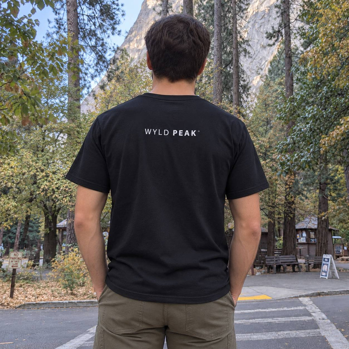A man viewed from behind wearing a black t-shirt with 'WYLD PEAK' printed in white on the back, standing outdoors in a park with trees and a mountain in the background.