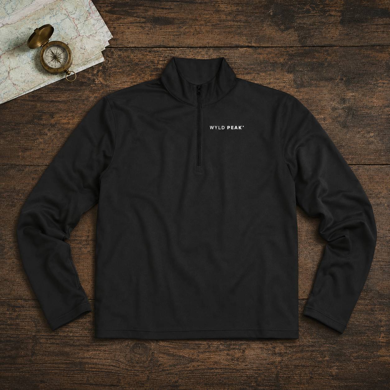 A black long-sleeve quarter-zip pullover with 'WYLD PEAK' printed in white on the left chest, laid flat on a rustic dark wood background with a map and compass prop in the top-left corner.