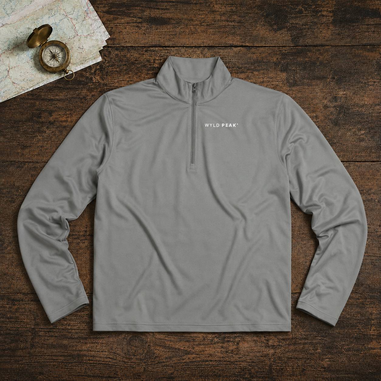 A light gray quarter-zip pullover with 'WYLD PEAK' printed in white on the left chest, laid flat on a dark wood background with a vintage map and compass as props.
