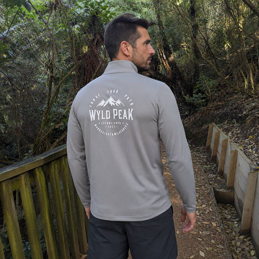 A man wearing a light gray long-sleeve quarter-zip pullover with a white 'WYLD PEAK' mountain logo on the back, standing on a wooden boardwalk in a forest.