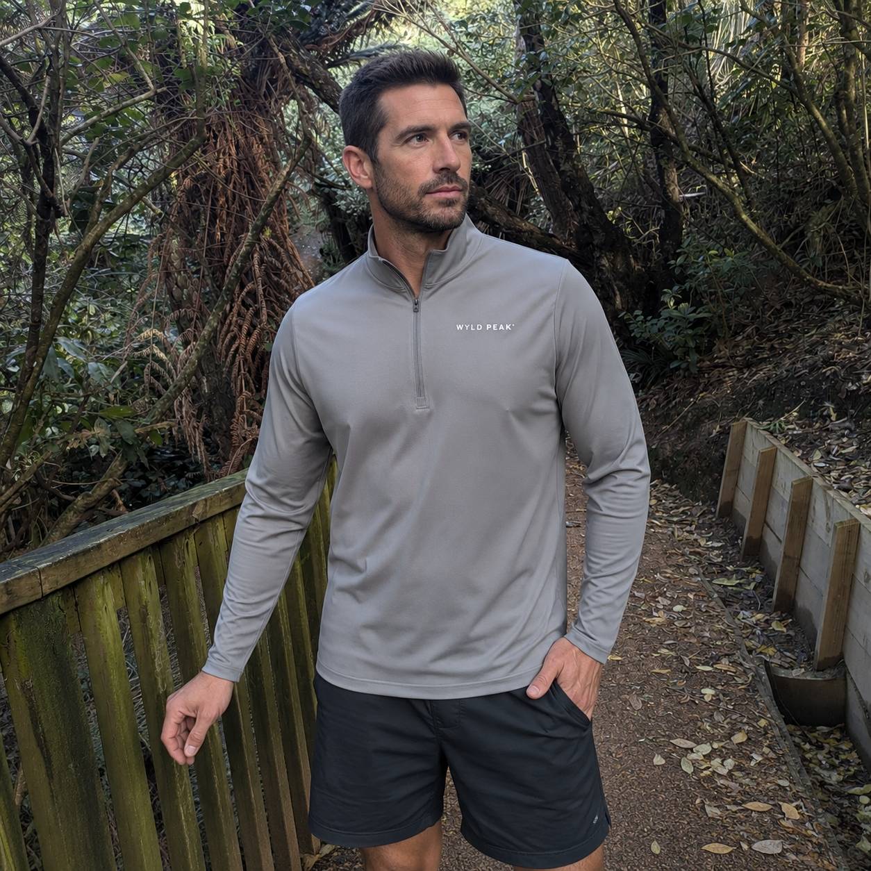 A man wearing a gray long-sleeve quarter-zip athletic shirt and black shorts standing on a forest path.