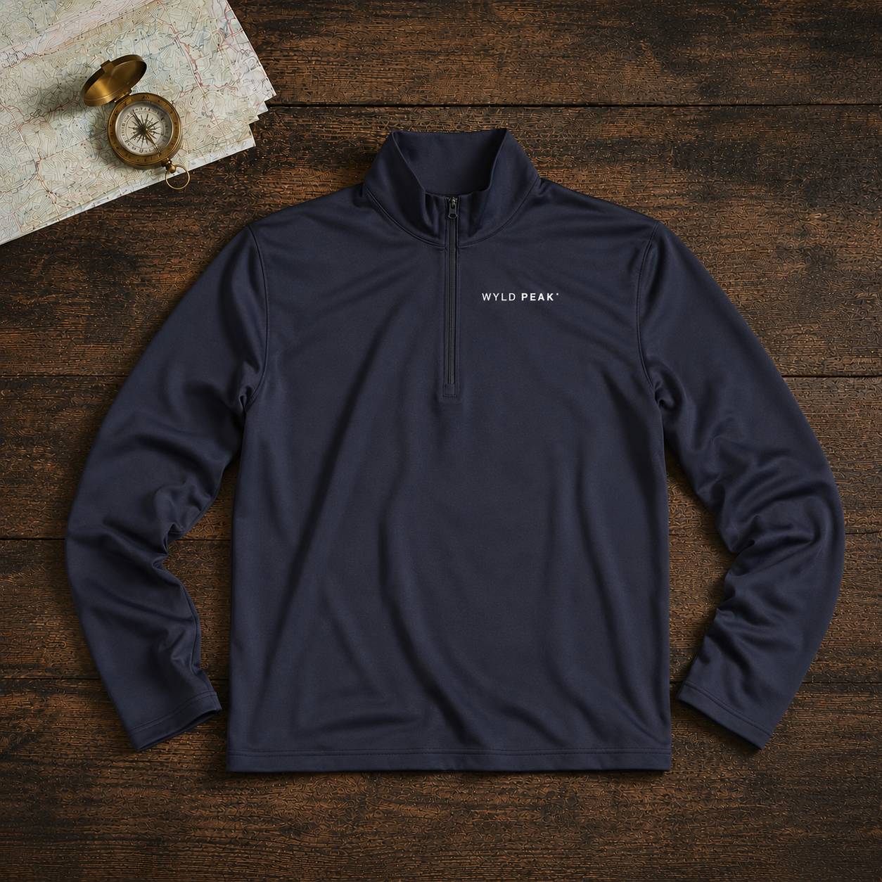 A navy blue quarter-zip pullover sweatshirt with 'WILD PEAK' printed in white on the left chest, laid flat on a dark wood background with a vintage compass and map as props.