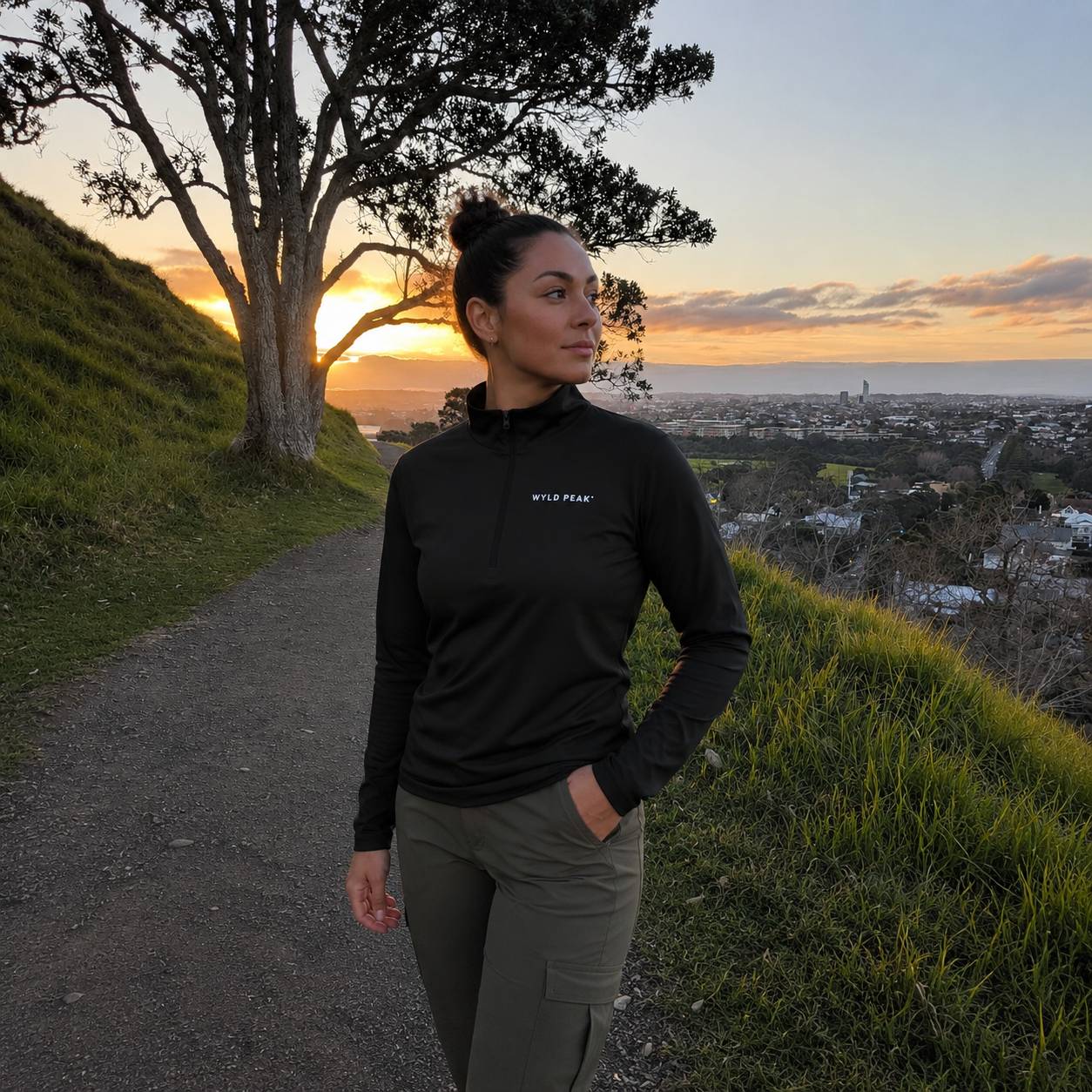 A woman stands on a hillside path at sunset wearing a black long-sleeve quarter-zip pullover with a small white 'WYLD PEAK' logo on the left chest, paired with olive cargo pants.