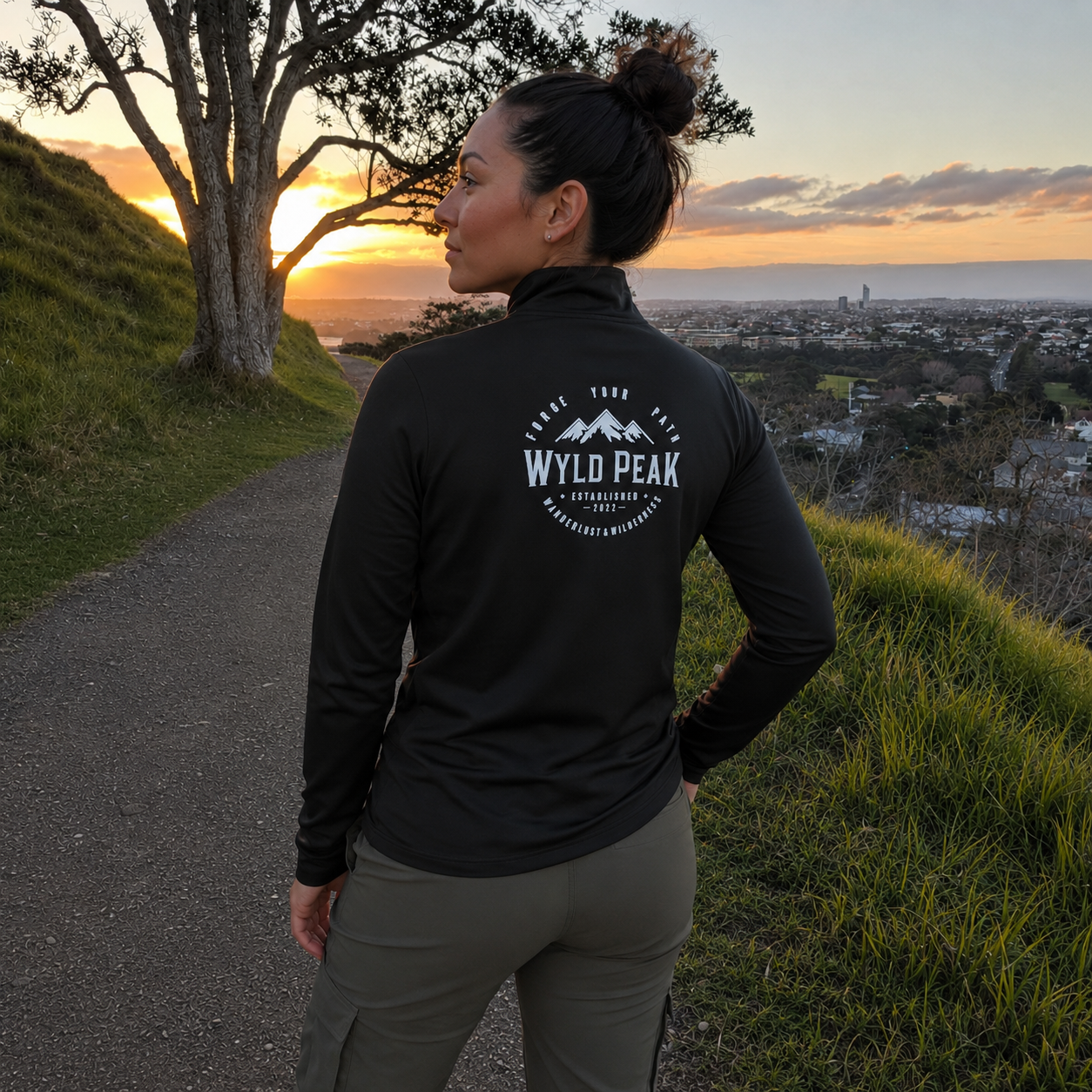 A woman stands on a hillside path at sunset wearing a black long-sleeve athletic top with a white circular 'WYLD PEAK' mountain logo on the back, paired with olive cargo pants, overlooking a scenic valley and ocean view.