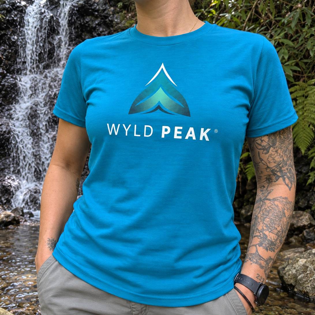 A person wearing a bright blue crewneck t-shirt with a 'WYLD PEAK' mountain logo printed on the chest, standing in front of a waterfall and green foliage.