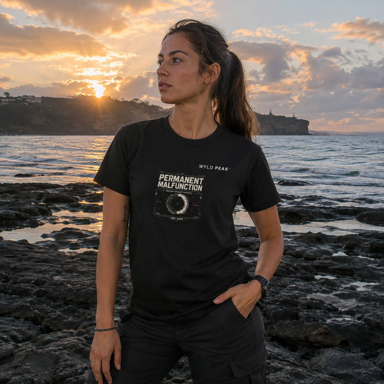 A woman wearing a black graphic t-shirt with 'WILD PEAK' and 'PERMANENT MALFUNCTION' printed on the front, standing on a rocky shoreline at sunset with the ocean and cliffs in the background.