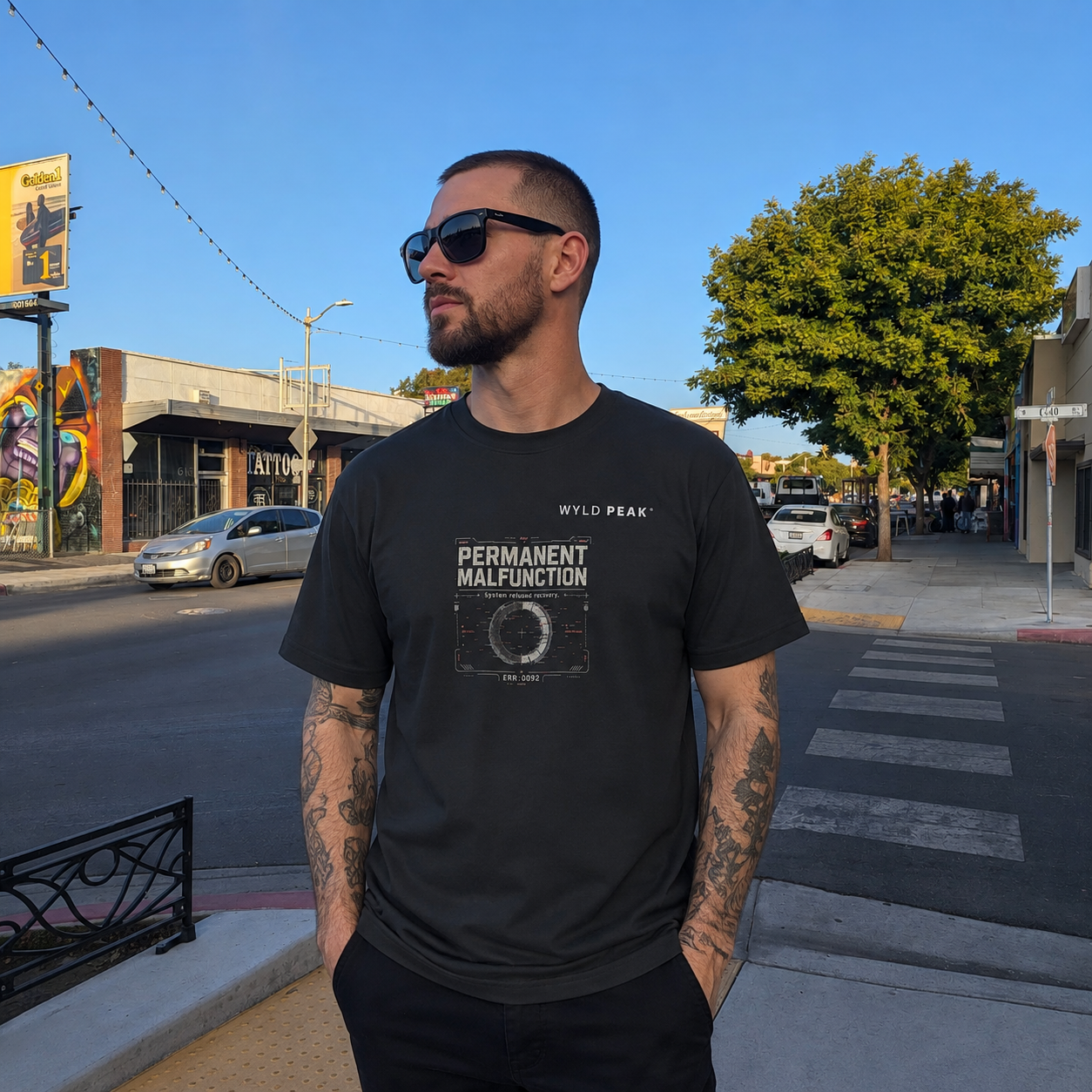 A man wearing a black graphic t-shirt with 'WYLD PEAK' and 'PERMANENT MALFUNCTION' printed on the front, standing on a city street corner.