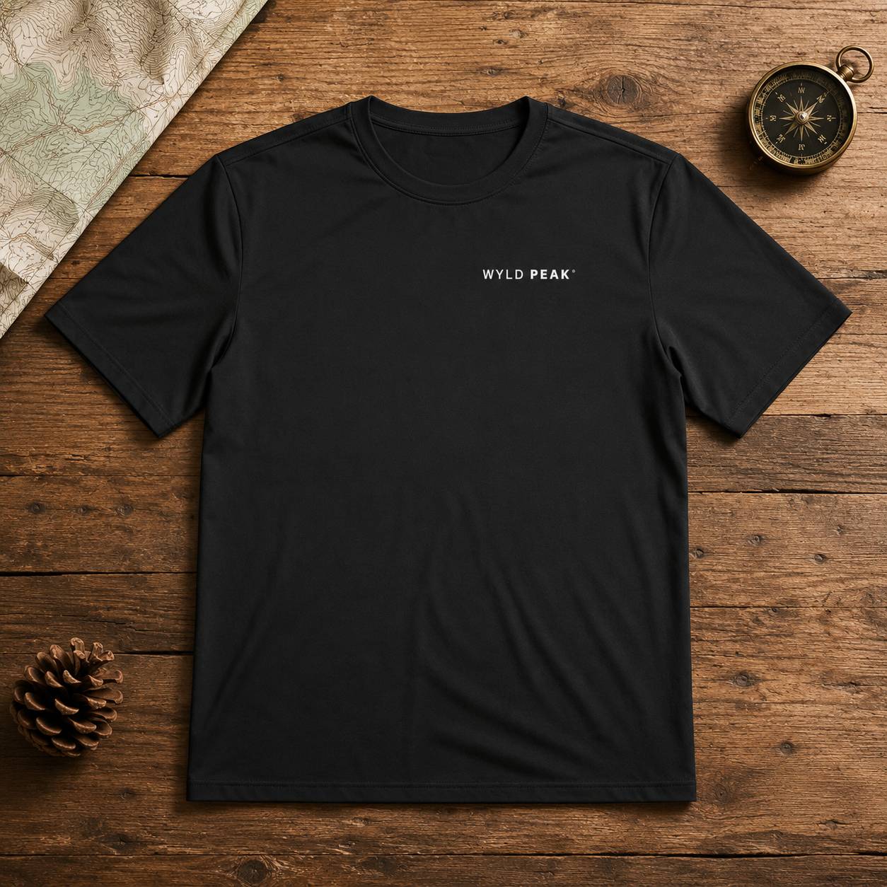 A black short-sleeve crewneck t-shirt with 'WYLD PEAK' printed in white on the left chest, laid flat on a rustic wooden surface with a pinecone, compass, and map as props.