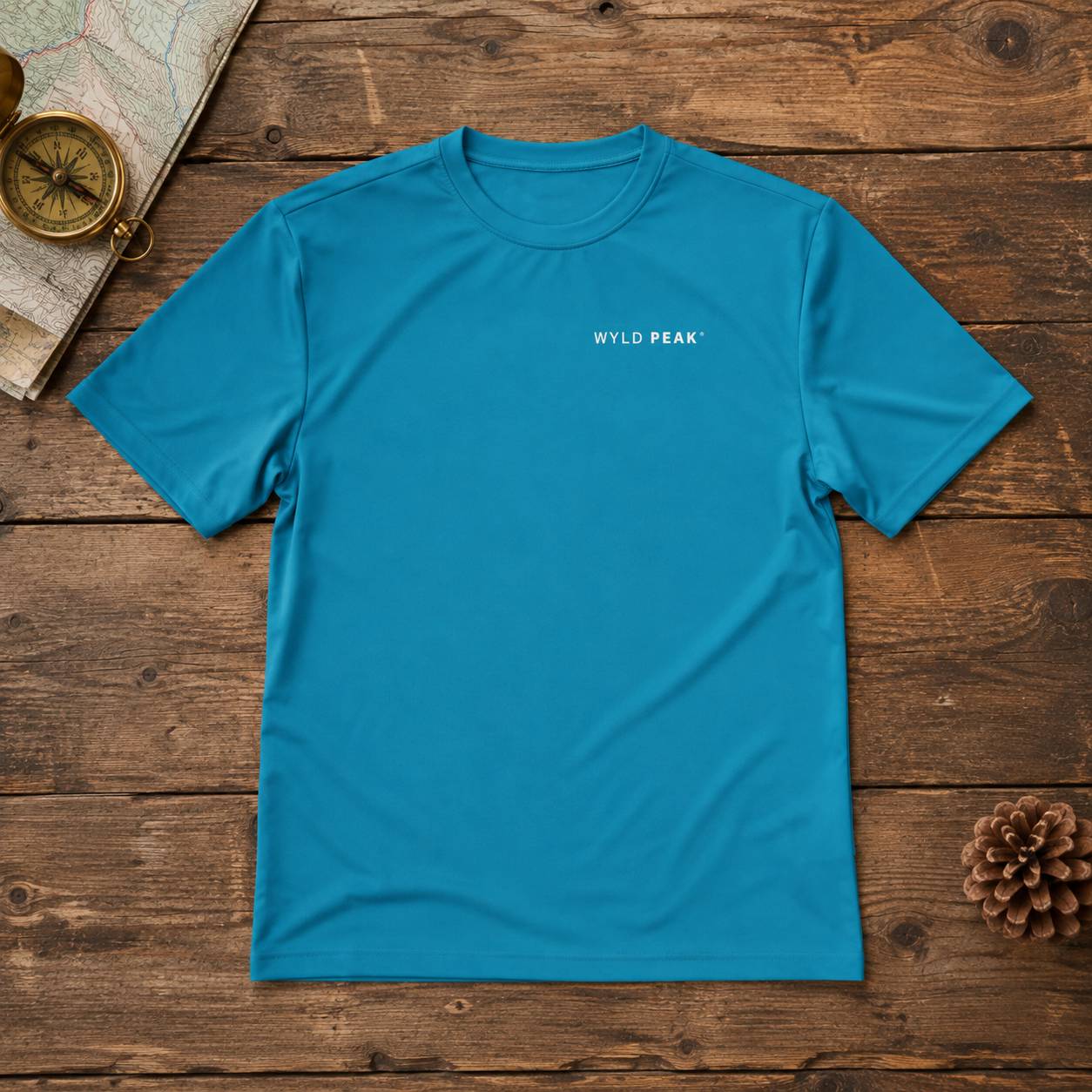 A bright blue crewneck t-shirt with 'WYLD PEAK' printed in white on the chest, laid flat on a rustic wooden background with a map, compass, and pinecone props.