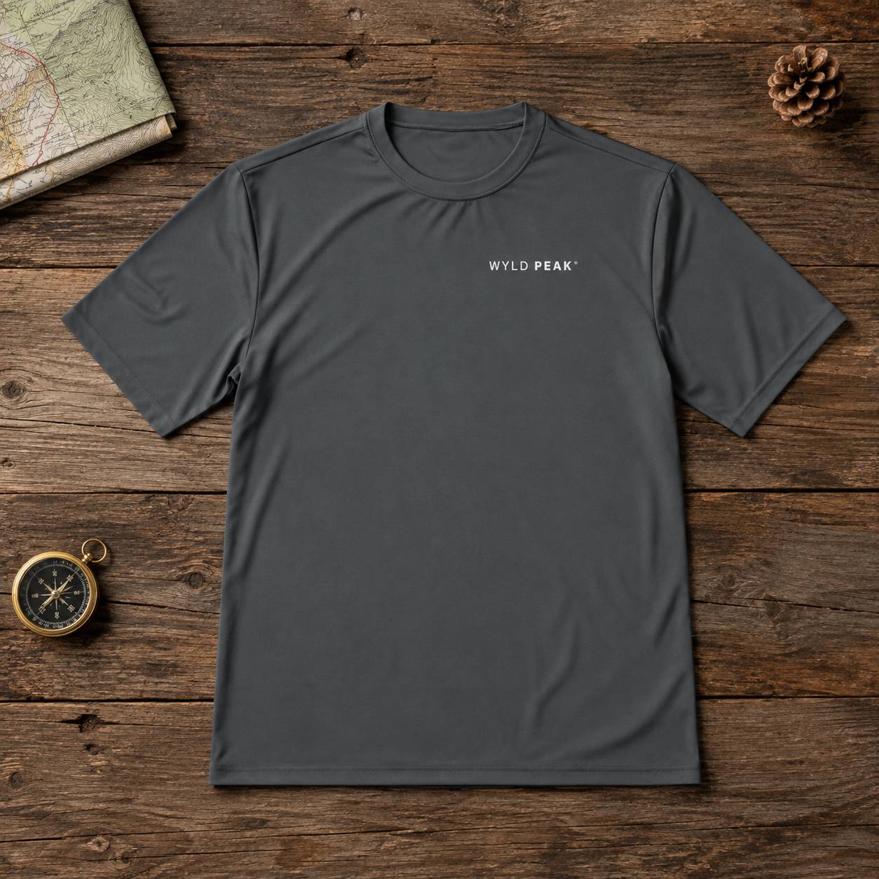 A dark gray crewneck t-shirt with 'WYLD PEAK' printed in white on the left chest, laid flat on a rustic wooden background with outdoor-themed props like a map, pinecone, and compass.