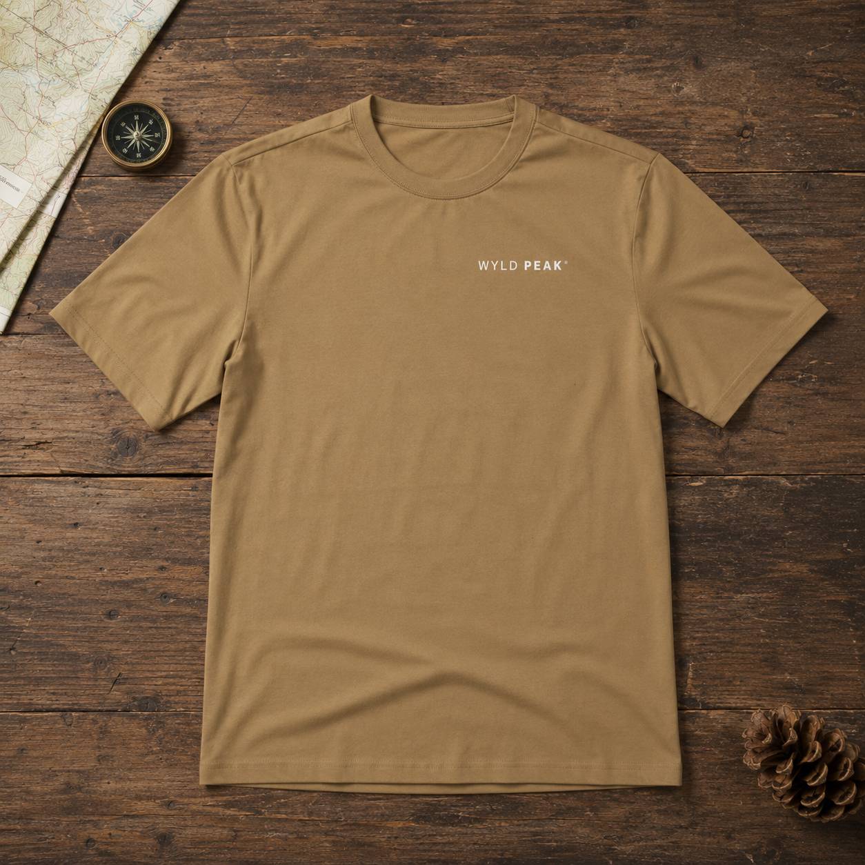 A tan crewneck t-shirt with 'WYLD PEAK' printed in white on the chest, laid flat on a rustic dark wood background with a map, compass, and pinecone props.