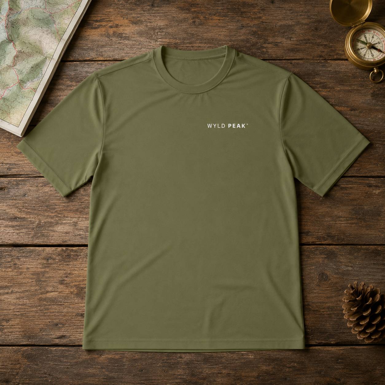 An olive green crewneck t-shirt with 'WYLD PEAK' printed in white on the chest, laid flat on a rustic wooden surface with a map, brass compass, and pinecone as props.