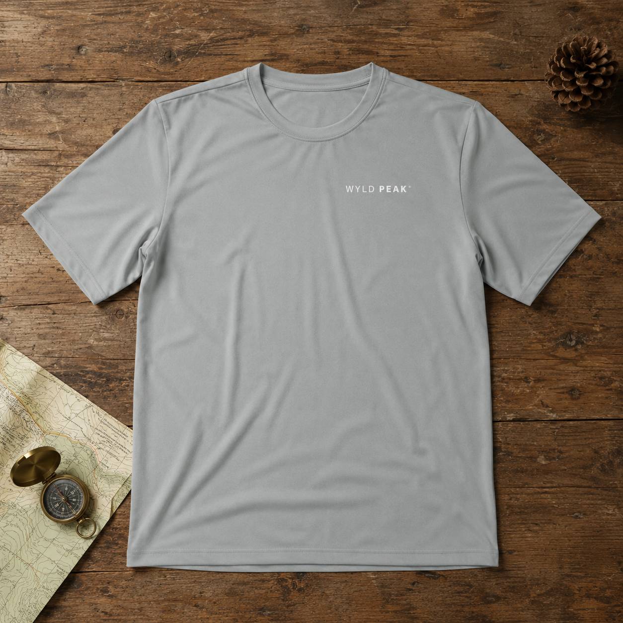 A light gray short-sleeve crewneck t-shirt with 'WILD PEAK' printed in white on the left chest, laid flat on a rustic wooden background with a pinecone and vintage map props.