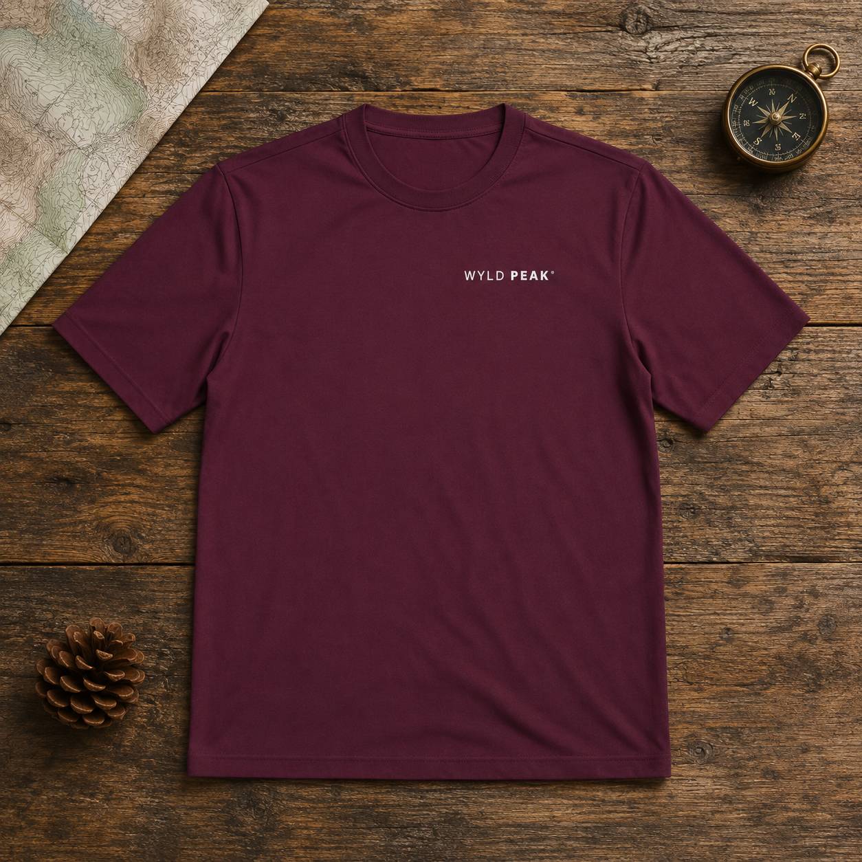 A maroon short-sleeve crewneck t-shirt with 'WYLD PEAK' printed in white on the left chest, laid flat on a rustic wooden surface with a pinecone, compass, and map as props.