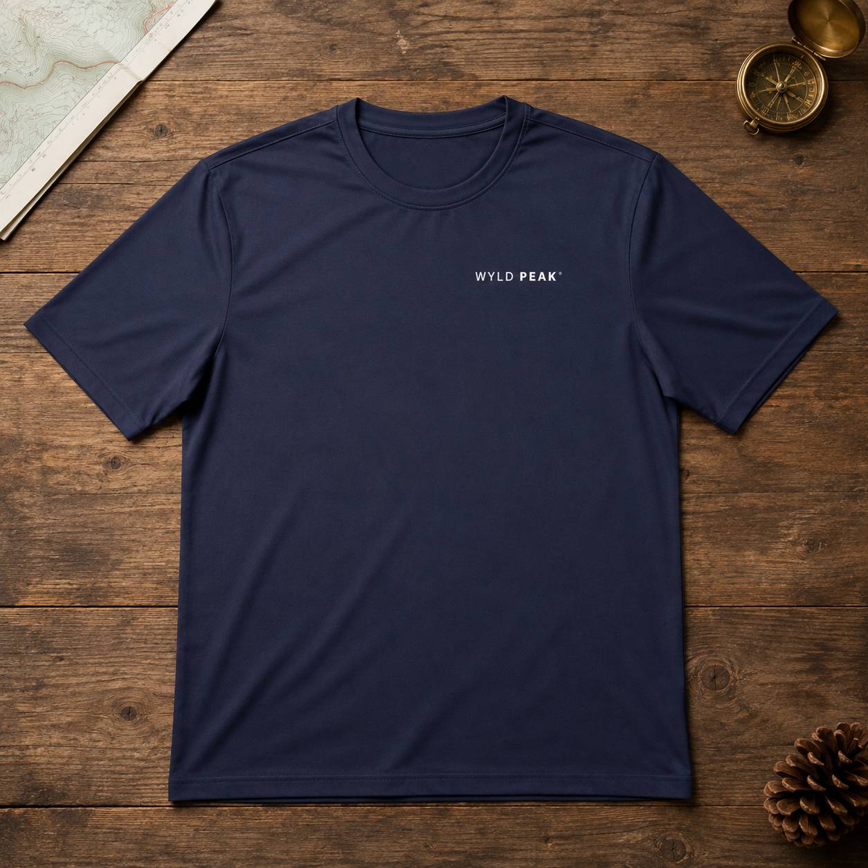 A navy blue short-sleeve crewneck t-shirt with 'WYLD PEAK' printed in white on the left chest, laid flat on a rustic wooden surface with a map, compass, and pinecone as props.