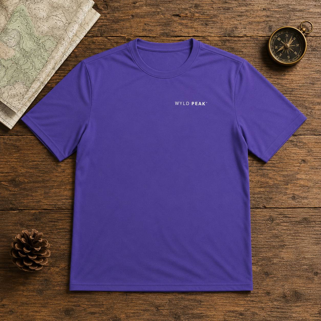 A purple short-sleeve crewneck t-shirt with 'WYLD PEAK' printed in white on the left chest, laid flat on a rustic wooden surface with a map, pinecone, and compass as props.