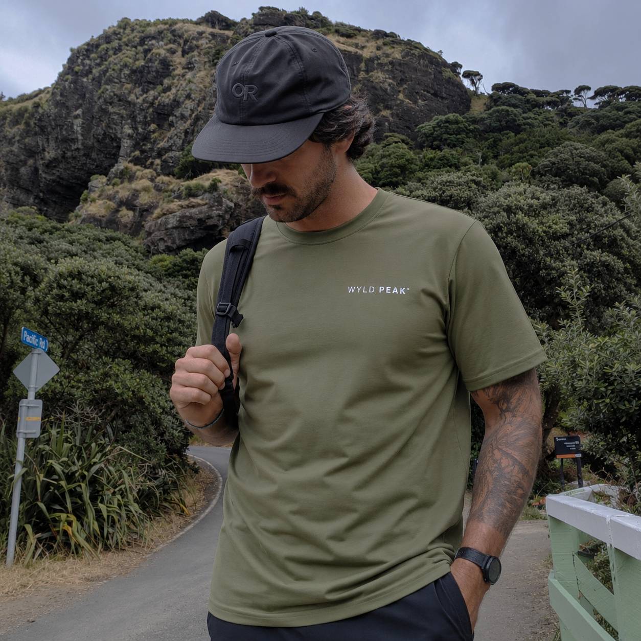 A man wearing an olive green t-shirt with 'WYLD PEAK' printed on the chest, a black baseball cap, and a black backpack strap, standing outdoors on a path with a rocky, forested hill in the background.