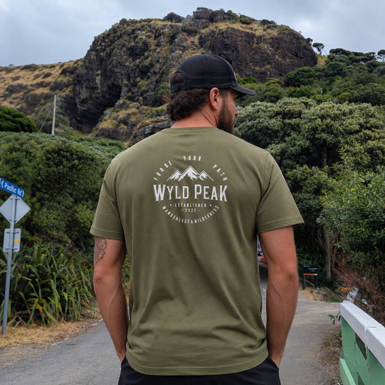 A man viewed from behind wearing an olive green t-shirt with a white 'Wyld Peak' mountain graphic on the back, standing on a path with a rocky hill and green forest in the background.