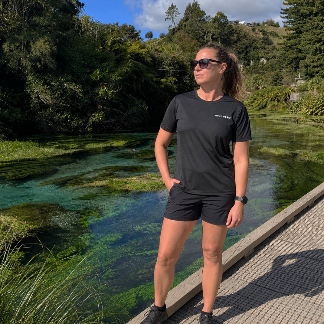 A woman wearing a black athletic t-shirt and matching shorts stands on a paved path next to a clear turquoise spring or stream, with lush green forest in the background. The brand name 'WYLD PEAK' is visible in white on the left chest of her shirt.