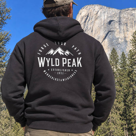 A man viewed from behind wearing a black hoodie with a white 'Wyld Peak' mountain graphic on the back, standing outdoors in front of a large rock formation and pine trees under a blue sky.