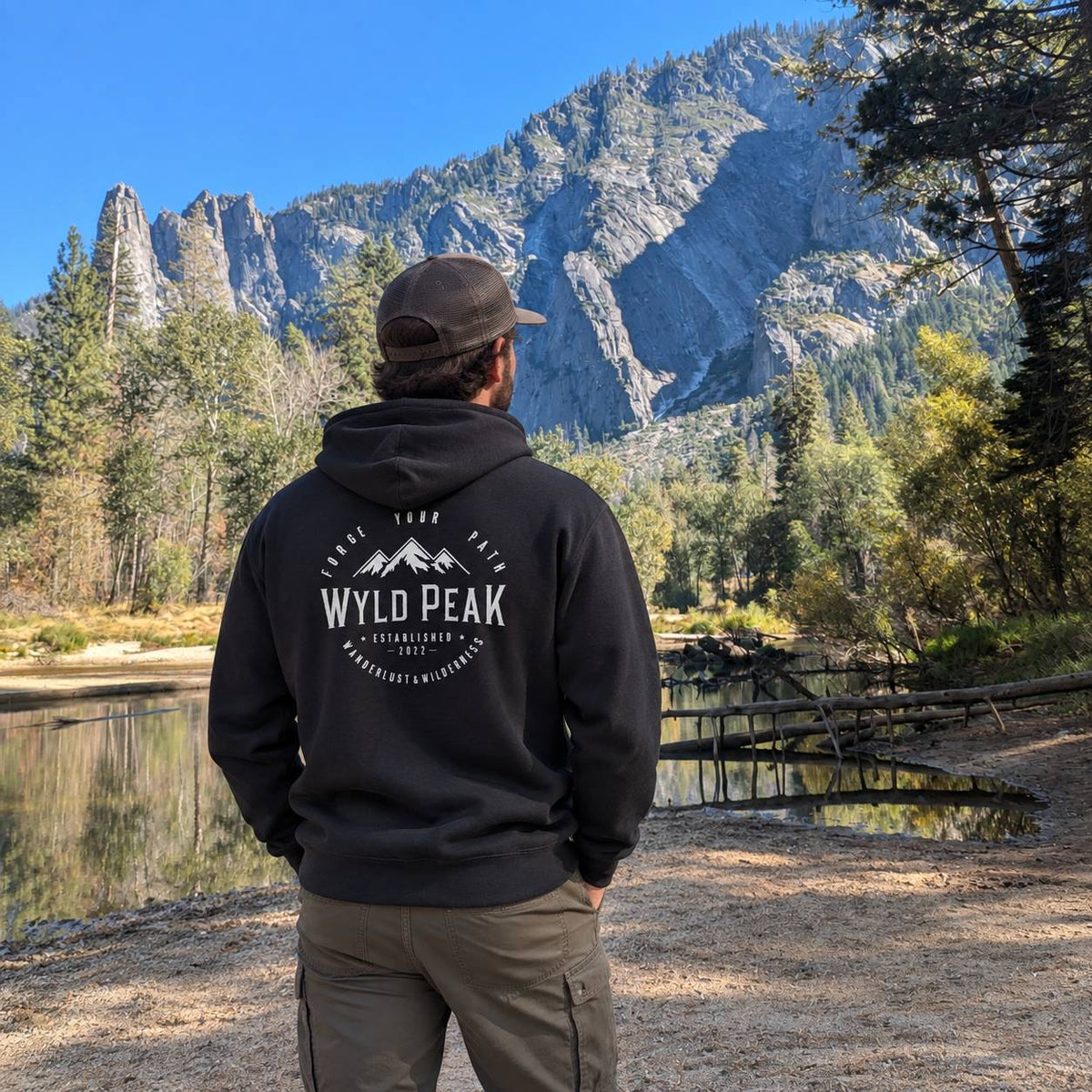 A man stands on a trail in a mountain landscape, viewed from behind, wearing a black hoodie with a white 'WYLD PEAK' graphic on the back and khaki pants. A lake, forest, and steep rocky mountains are visible in the background under a blue sky.