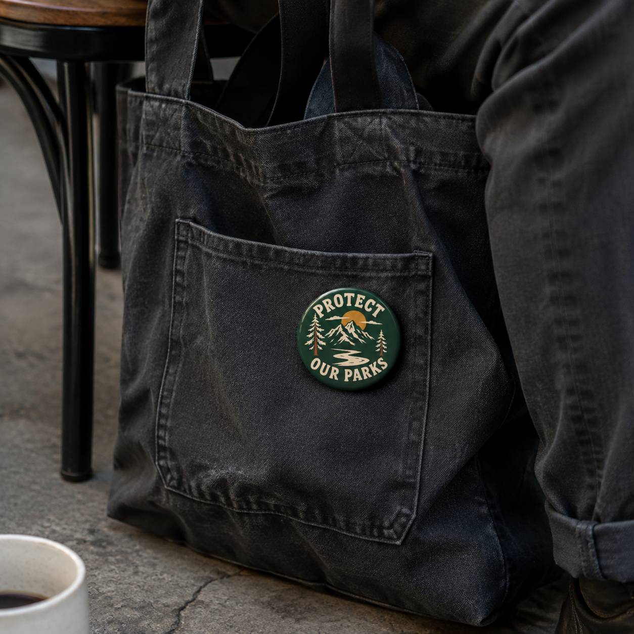 A dark charcoal gray canvas tote bag with a front pocket, featuring a round green and gold 'PROTECT OUR PARKS' pin badge on the pocket. The bag is shown in a lifestyle setting next to a person wearing dark gray pants, with a coffee cup and table visible in the background.