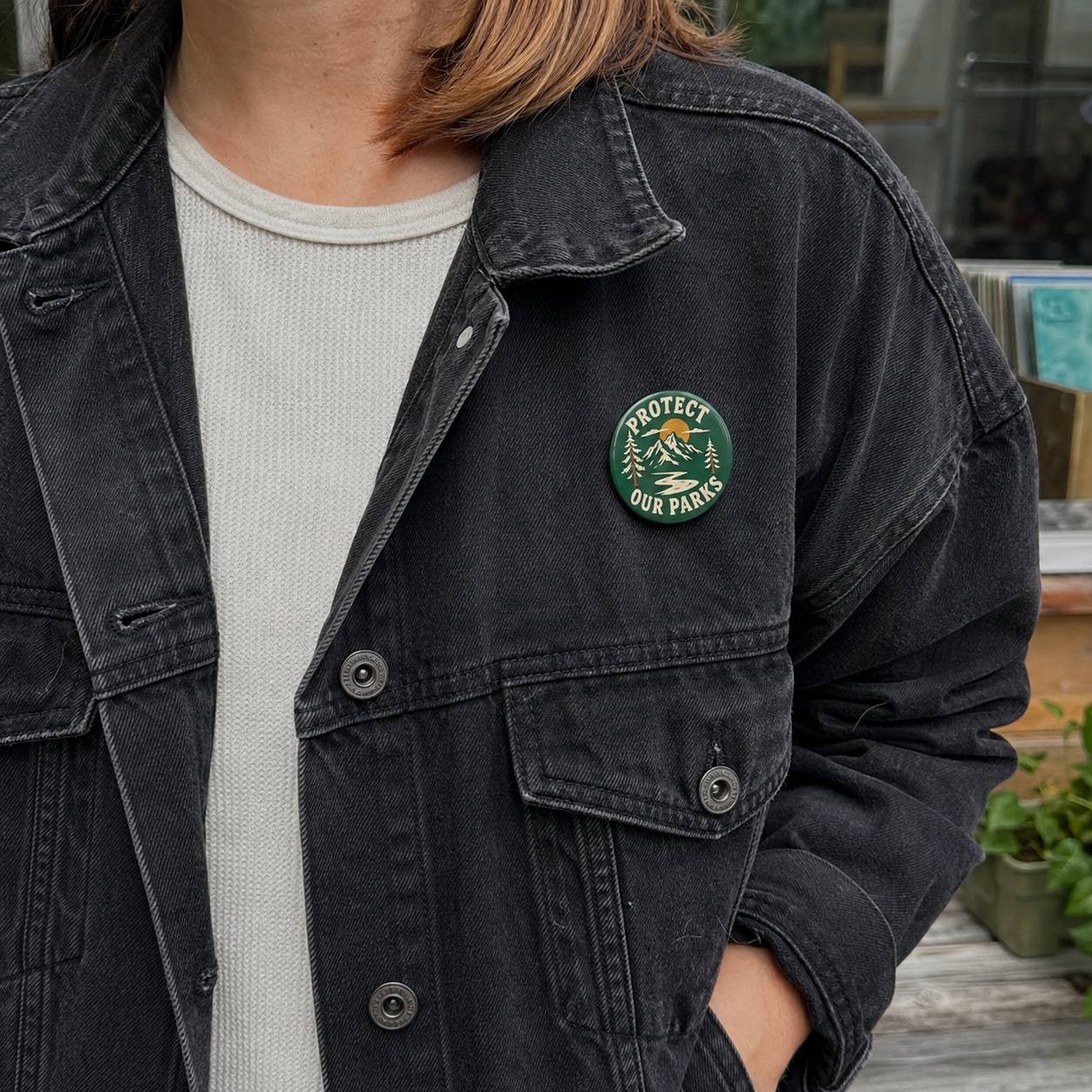 A person wearing a washed black denim jacket over a light gray ribbed t-shirt. The jacket features a circular green and gold embroidered patch on the left chest that reads 'PROTECT OUR PETS'.