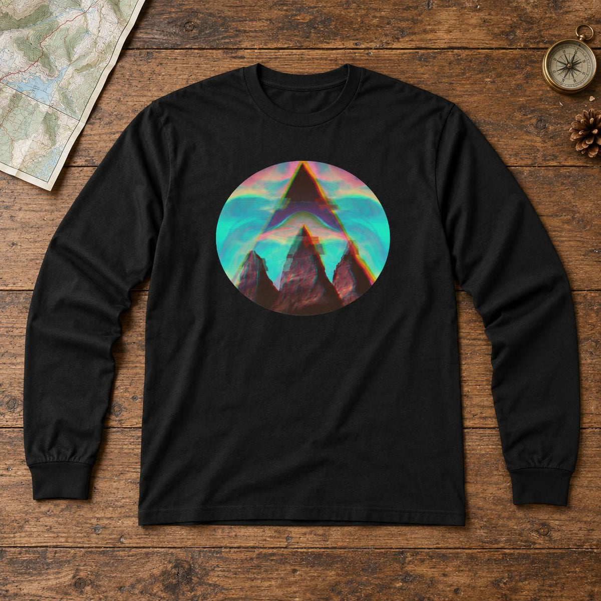 A black long-sleeve t-shirt with a circular graphic print of a mountain peak in front of a colorful teal and pink sky, laid flat on a rustic wooden background with a map, compass, and pinecone props.