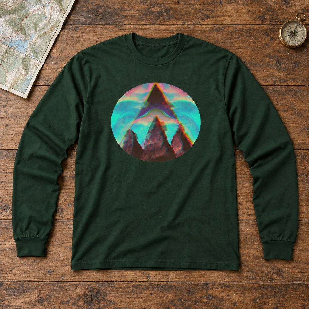 A dark forest green long-sleeve t-shirt with a circular graphic print of a mountain peak in front of a colorful sunset sky, laid flat on a rustic wooden background with a map and compass props.