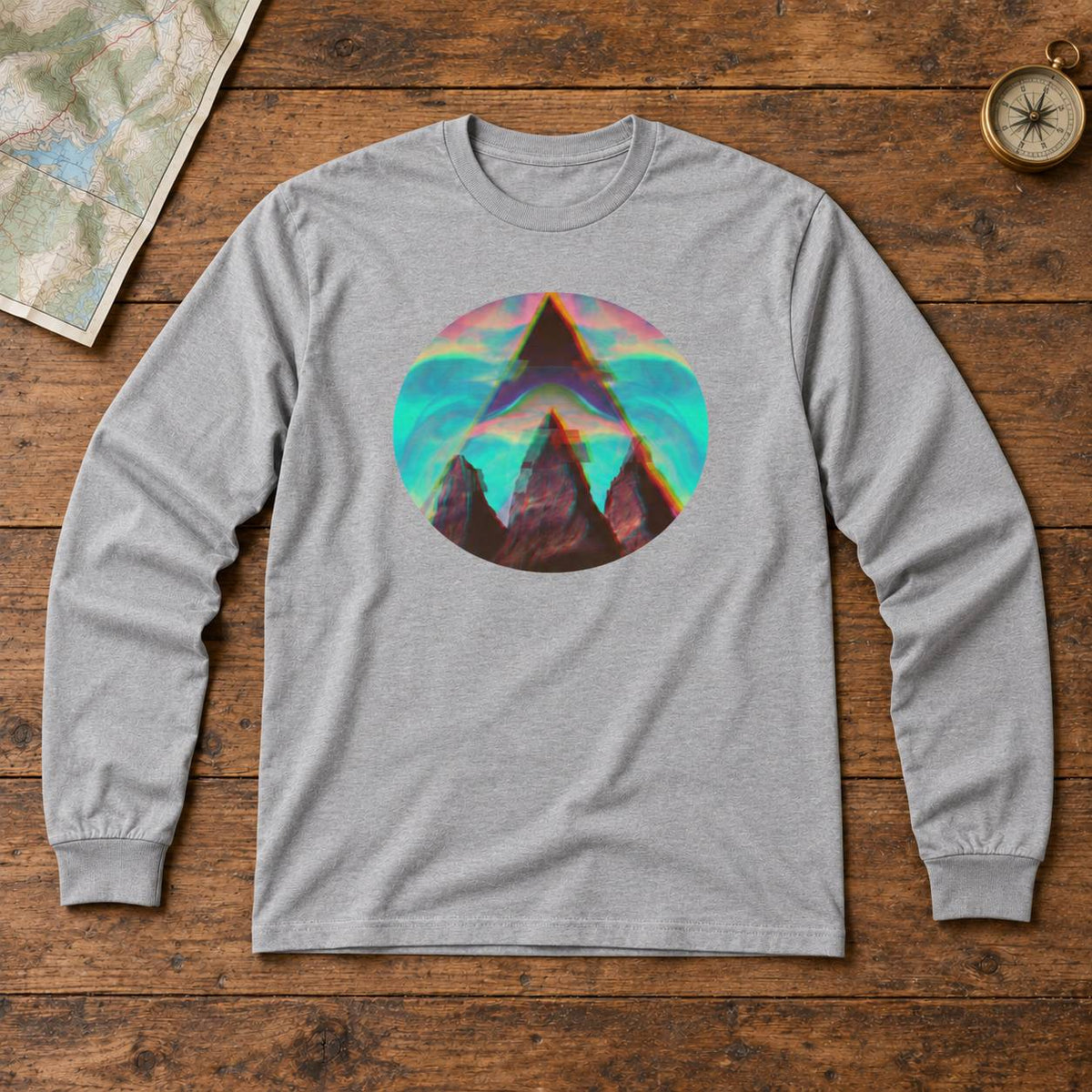 A heather gray long-sleeve t-shirt with a colorful retro circular graphic of a mountain peak, laid flat on a rustic wooden background with a map and compass props.