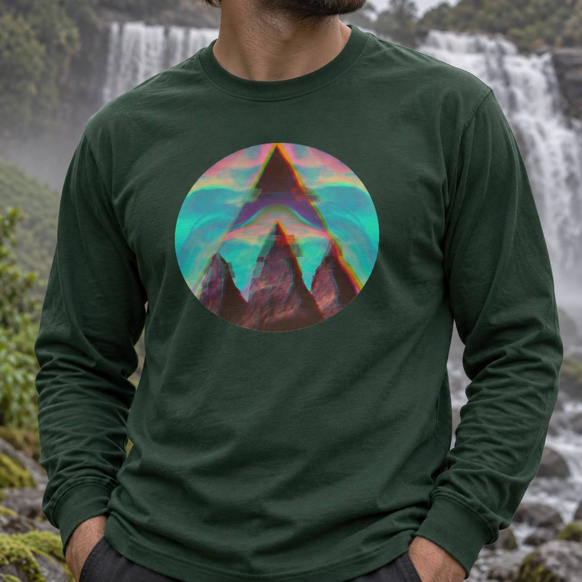 A man wearing a dark forest green long-sleeve t-shirt with a colorful circular graphic of a mountain peak on the chest, set against a blurred background of waterfalls and greenery.
