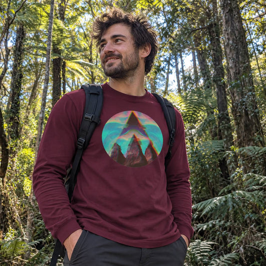 A man wearing a burgundy long-sleeve t-shirt with a colorful circular mountain graphic on the chest, standing in a lush green forest.